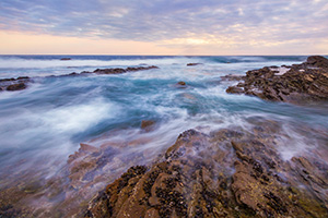 Waves wash around the rocks on the beach at Crystal Cove State Park, California while the sun sets in the distance. - California Landscape Photograph