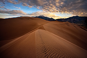 On a cool, autumn morning, silence reigned across the scenic Great Sand Dunes National Park, the quiet only occasionally broken by the sound of a breeze flowing through the dunes.  On top of one of the larger dunes, I captured the patterns of the sand as the dunes drift into one another, creating a path to the distant Sangre de Cristo Mountains.   Beyond the far peaks, the rising sun illuminates the cloud bank with a yellowish-orange tint. - Colorado Photograph