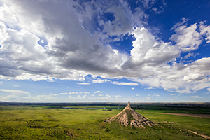 From a vantage point on a nearby bluff Chimney Rock glows in the light of the warm afternoon sun under a dark blue sky filled with clouds. - Nebraska Photograph