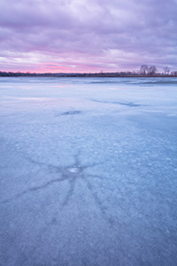 On this evening at DeSoto National Wildlife Refuge a quiet stillness prevailed.  I wandered the shore of the lake looking for interesting patterns in the now melting ice.  I found some interesting star shapes that had emerged from the recent thaws.  Sunset was near and although it was cloudy, I was hoping for a little illumination of the clouds.  After a while I gave up and begin leaving when a bit of sun broke free and I quickly set up again and captured this image. - Nebraska Landscape Photograph