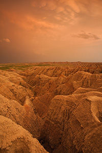 Orange hues from the last light of the setting sun illuminate storm clouds as they pass over Badlands National Park in South Dakota, dark clouds contrast with the desolate landscape. - South Dakota Photograph