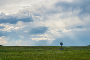 The sun peeks through a group of storm clouds illuminating a windmill deep within the Sandhills of Nebraska as storm clouds roll in the distance. - Nebraska Photograph